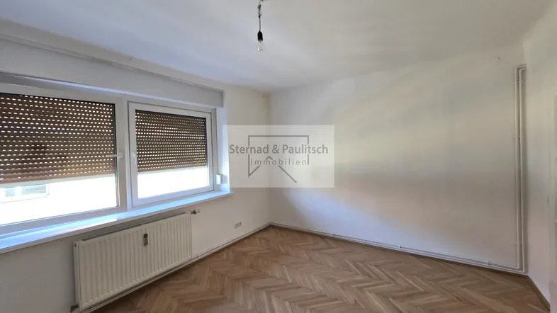 Helles Zimmer mit Fischgrätparkettboden und Fenster mit Jalousien, ideal als Wohn- oder Schlafzimmer.