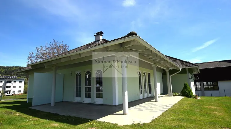 Modernes Einfamilienhaus mit überdachter Terrasse und großem Garten unter blauem Himmel.