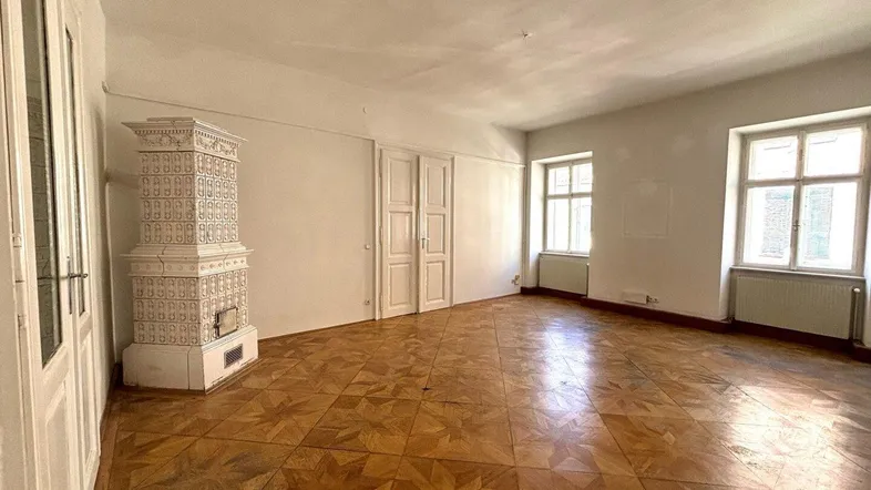 Geräumiges Wohnzimmer mit traditionellem Kachelofen und Fischgrätparkett, hell durch große Fenster.