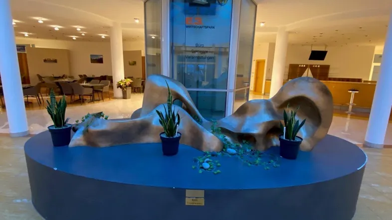 Geräumige Lobby mit moderner Skulptur, Glasaufzug und Sitzbereichen im Hintergrund.