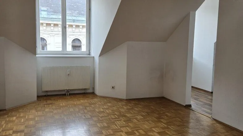 Helles Zimmer mit Dachschrägen, Parkettboden und Fensterblick auf die umliegenden Gebäude.