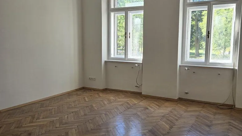 Helles, leeres Zimmer mit klassischem Fischgrätparkett und zwei großen Fenstern, die viel Licht hereinlassen.