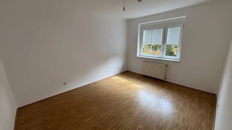 Helles, leeres Zimmer mit Parkettboden und Fenster, das viel Tageslicht hereinlässt.