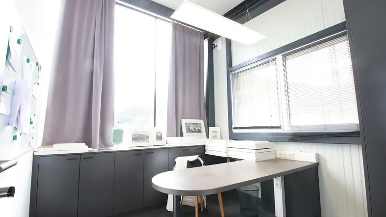 Modernes Büro mit großen Fenstern, dunklen Vorhängen und einem ovalen Schreibtisch, ideal für konzentriertes Arbeiten.