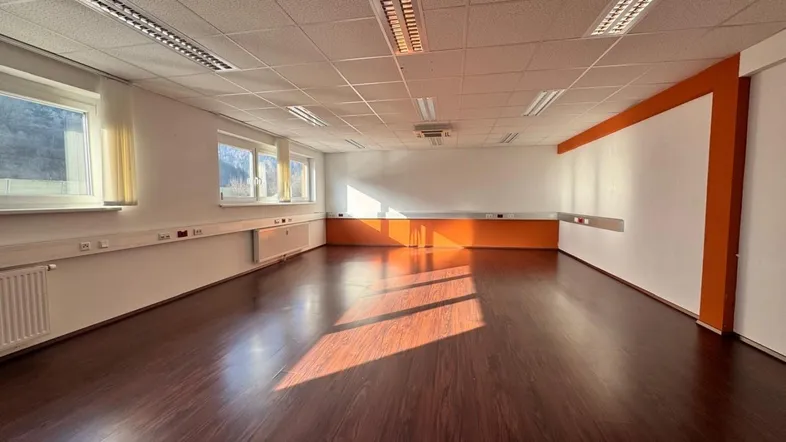 Geräumiger, leerer Büroraum mit dunklem Laminatboden, weißen Wänden und großen Fenstern, die viel Tageslicht hereinlassen.