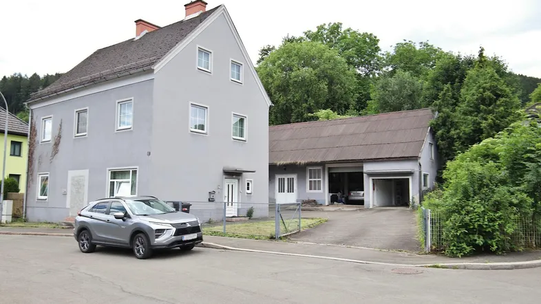 Graues Wohnhaus mit angrenzendem Nebengebäude und einem geparkten Auto davor.