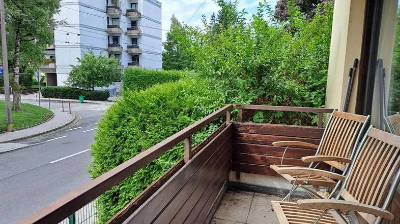 Gemütlicher Balkon mit Holzboden und Sitzgelegenheiten, umgeben von grünen Hecken und Blick auf die Straße.