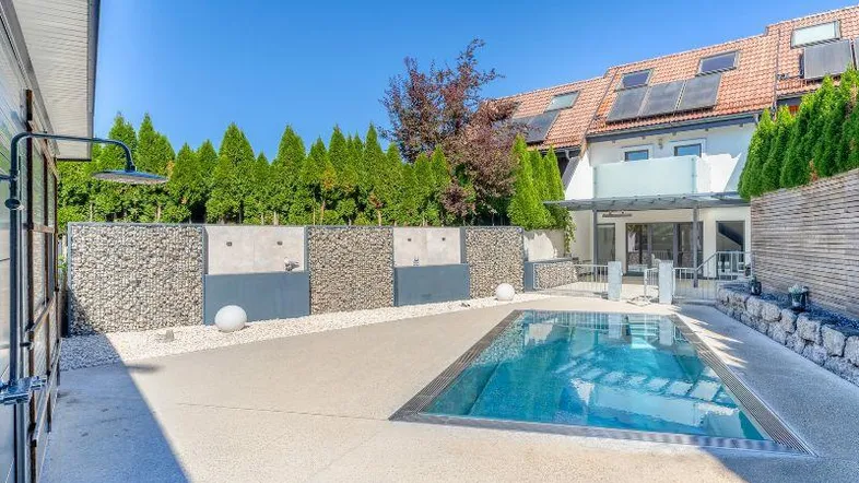 Moderner Außenbereich mit Pool, Außendusche und gepflegter Bepflanzung, umgeben von einer Gabionenmauer.