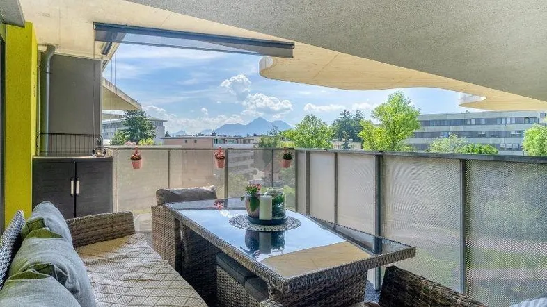 Möblierter Balkon mit Rattanmöbeln, Glasgeländer und weitem Blick auf die Berge und Umgebung.