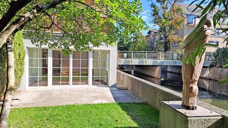 Sonnige Terrasse mit angrenzendem Rasen und Blick auf den Kanal, umgeben von Bäumen.