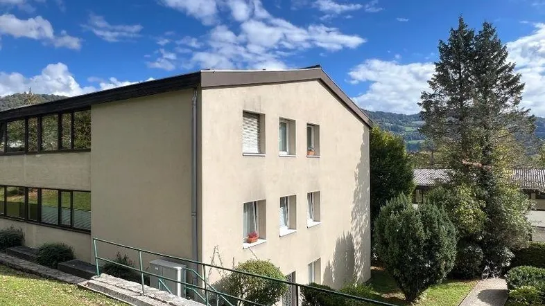 Ansicht des Mehrfamilienhauses mit heller Fassade und umliegender Grünfläche unter blauem Himmel.