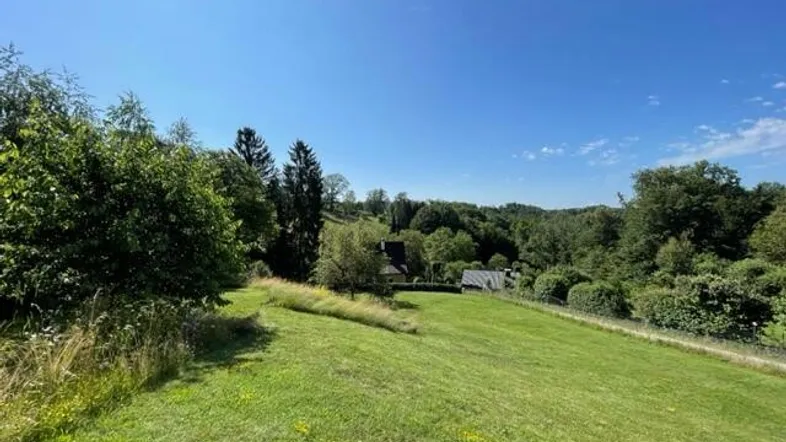 Weitläufige grüne Landschaft mit sanften Hügeln und vereinzelten Häusern unter blauem Himmel.