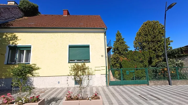 Gelbes Einfamilienhaus mit grünen Fensterläden und gepflastertem Vorgarten unter blauem Himmel.