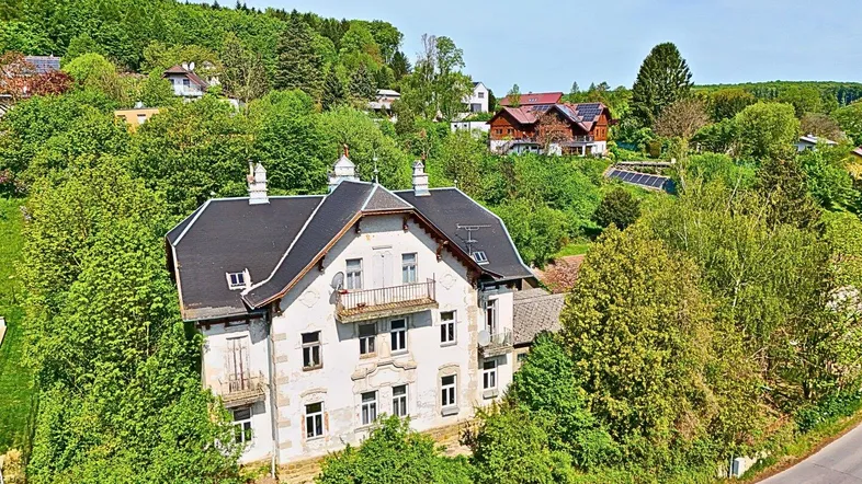 Historisches Gebäude mit Mansarddach, umgeben von üppigem Grün und Bäumen in ländlicher Umgebung.