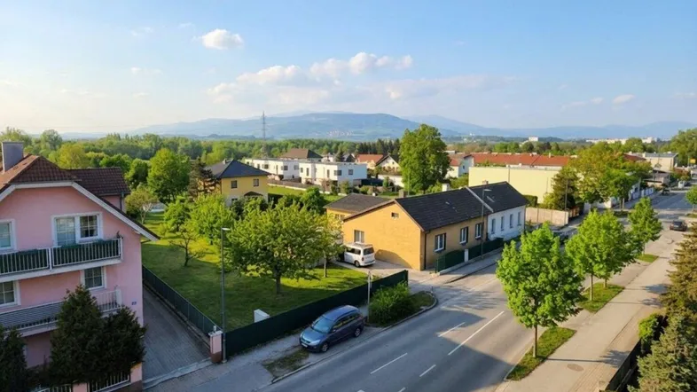 Luftaufnahme einer Wohngegend mit Häusern, Bäumen und Bergen im Hintergrund unter blauem Himmel.