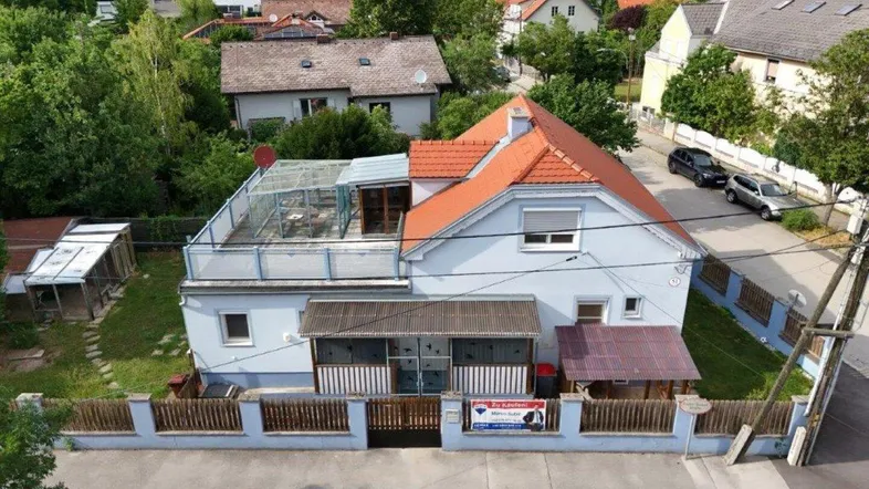 Luftaufnahme eines Einfamilienhauses mit hellblauer Fassade, rotem Ziegeldach, Garten und Bergblick.