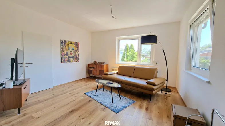 Helles Wohnzimmer mit Holzboden, beigem Sofa und Blick ins Grüne durch große Fenster.