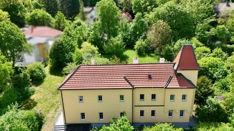 Historisches Gebäude mit gelber Fassade und rotem Ziegeldach, umgeben von üppigem Grün.