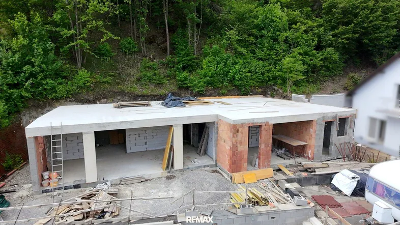 Rohbau des Erdgeschosses mit Betonplatten und Ziegelwänden, umgeben von viel Grün.