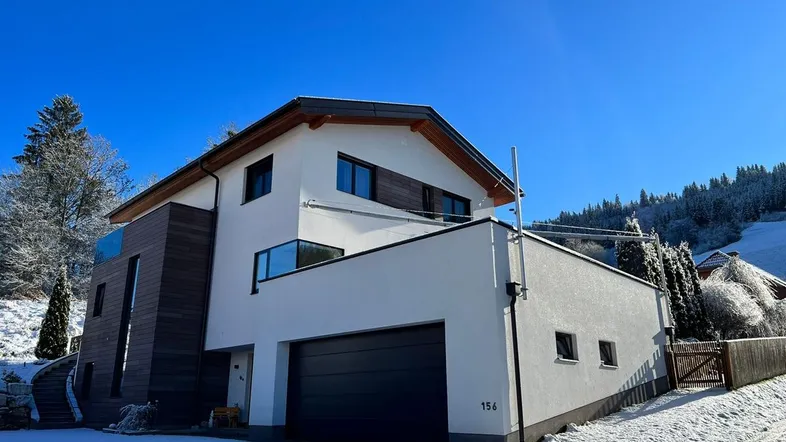 Modernes Einfamilienhaus mit weißer Fassade und Holzverkleidung, umgeben von Schnee und Bäumen.