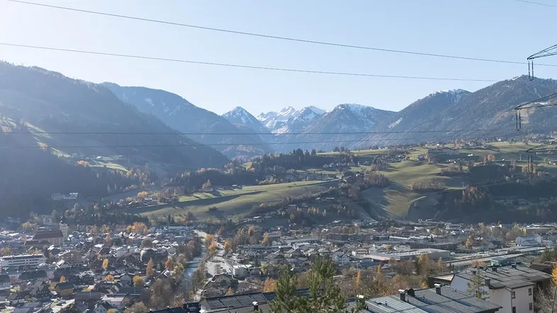 Panoramablick über ein Tal mit einer Stadt, umgeben von bewaldeten Hügeln und schneebedeckten Bergen.