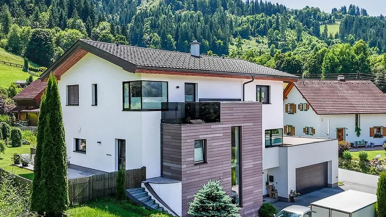 Modernes Einfamilienhaus mit weißer Fassade, großen Fenstern und Bergblick in grüner Umgebung.
