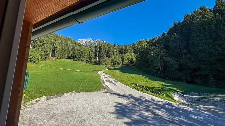 Blick von einer überdachten Loggia auf eine grüne Wiese, einen Wald und majestätische Berge unter blauem Himmel.