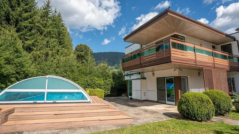 Außenansicht des Hauses mit überdachtem Poolbereich und Holzterrasse, umgeben von Bäumen.