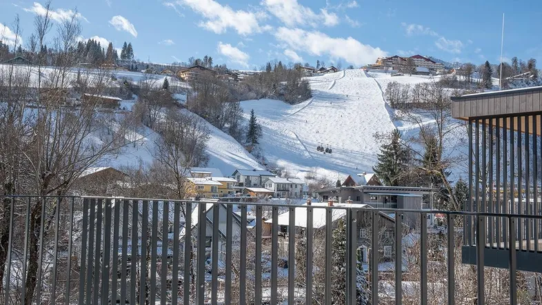 Blick vom Balkon auf die verschneite Berglandschaft mit Skipiste und umliegenden Häusern.