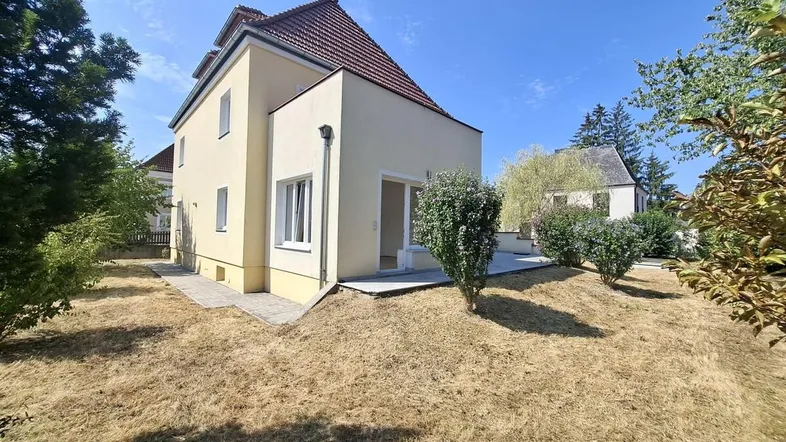 Hausfassade mit trockenem Rasen und Bäumen unter blauem Himmel an einem sonnigen Tag.