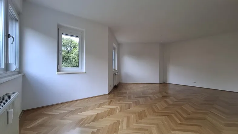 Lichtdurchflutetes Wohnzimmer mit Fischgrätparkett und mehreren Fenstern, die viel Tageslicht hereinlassen.