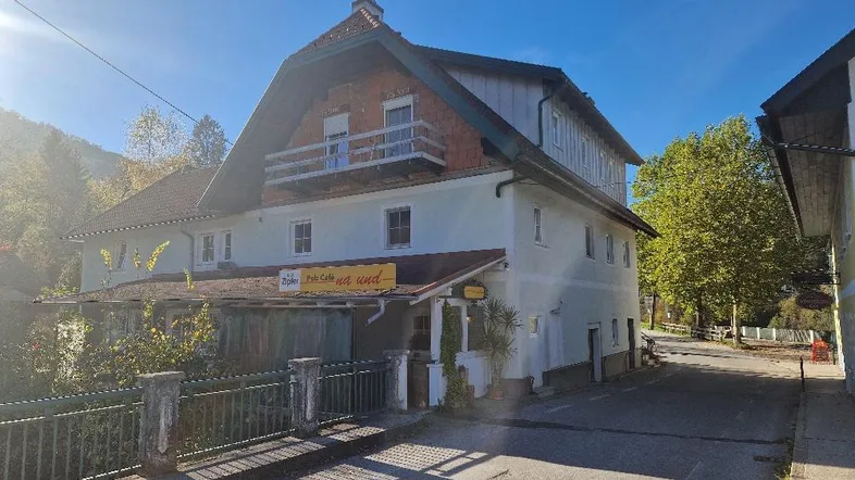 Traditionelles Gebäude mit Satteldach und Balkon, umgeben von grüner Natur und einer Straße.