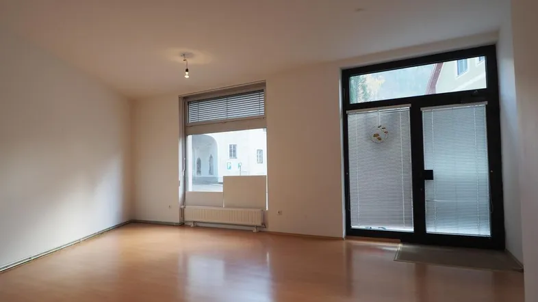 Heller, leerer Raum mit Laminatboden und großen Fenstern, die einen Blick auf einen Innenhof bieten.
