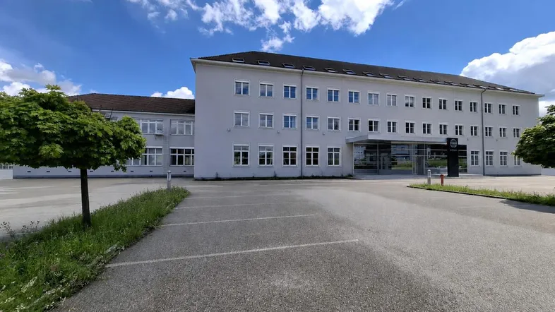 Großes Bürogebäude mit mehreren Stockwerken und einem geräumigen Parkplatz davor unter blauem Himmel.