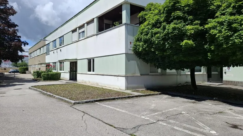 Gebäudefassade mit Parkplätzen und Bäumen unter einem bewölkten Himmel.