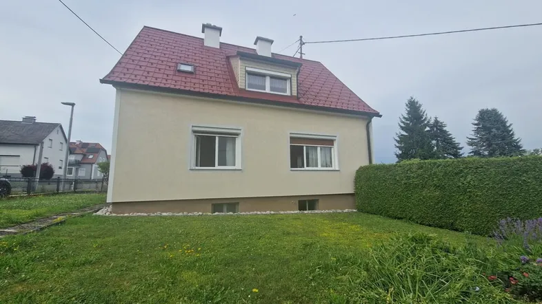 Hausfassade mit rotem Ziegeldach und gepflegtem Vorgarten unter bewölktem Himmel.