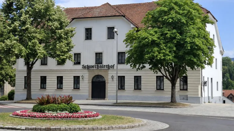 Traditionelles Gebäude "Schwechaterhof" mit weißer Fassade, umgeben von Bäumen und einem Blumenbeet.