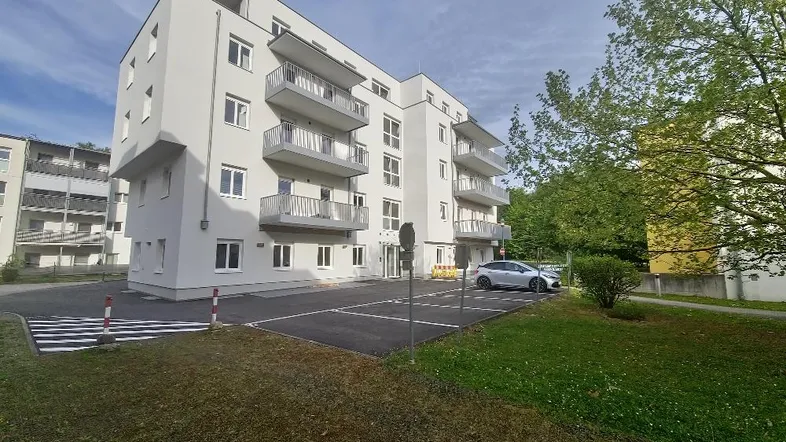 Modernes Mehrfamilienhaus mit mehreren Balkonen und Parkplätzen im Freien, umgeben von Grünflächen.