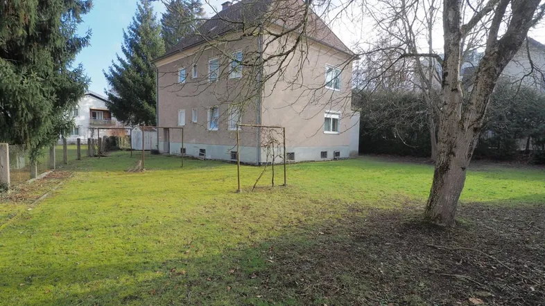 Großes Haus mit weitläufigem Rasenbereich und altem Baumbestand im Vordergrund.