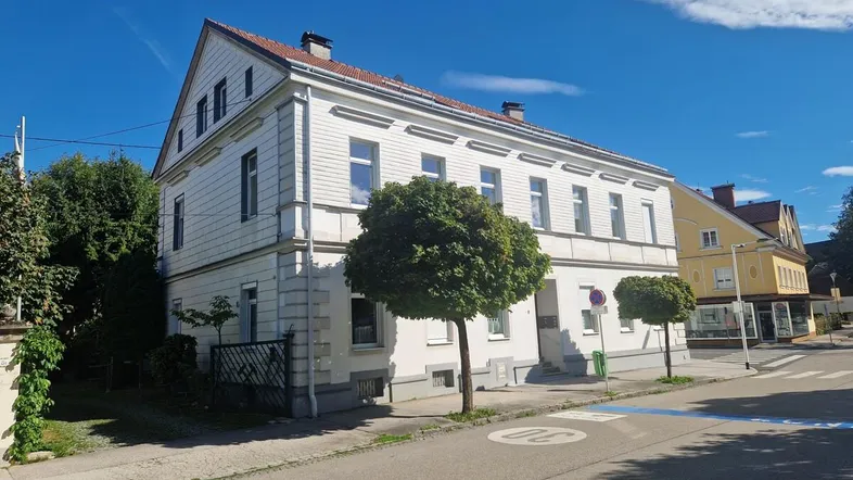 Weißes Mehrfamilienhaus mit Giebeldach an einer ruhigen Straße mit Bäumen und blauem Himmel.