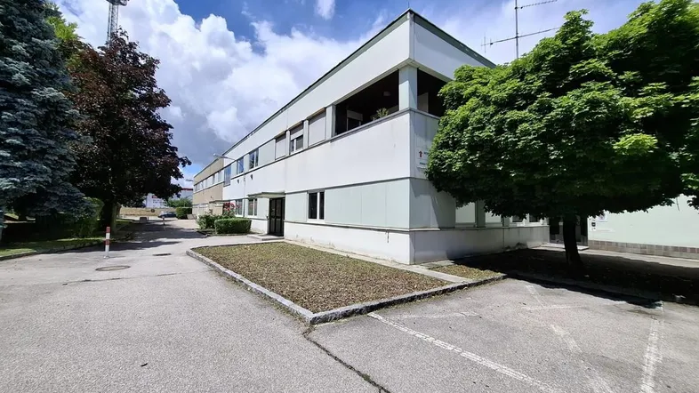 Gebäudeaußenansicht mit Parkplätzen und Bäumen unter bewölktem Himmel, zeigt funktionale Architektur.