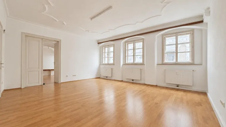 Geräumiges Zimmer mit hellem Parkettboden, Stuckdecke und drei Fenstern, ideal als Wohnbereich.