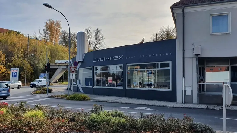Gebäude an einer Straßenecke mit großen Fenstern und umliegender Vegetation unter bewölktem Himmel.