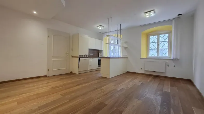 Heller offener Wohnbereich mit integrierter Küche, Holzboden und einem Fenster mit Gitter, das Tageslicht spendet.