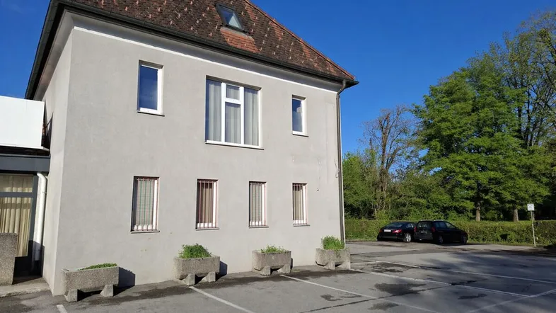 Graues Gebäude mit rotem Ziegeldach und Parkplätzen davor unter blauem Himmel.