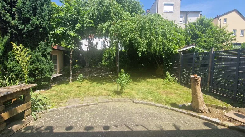 Grüner Gartenbereich mit Rasenfläche, Bäumen und einer kleinen gepflasterten Terrasse im Sonnenschein.