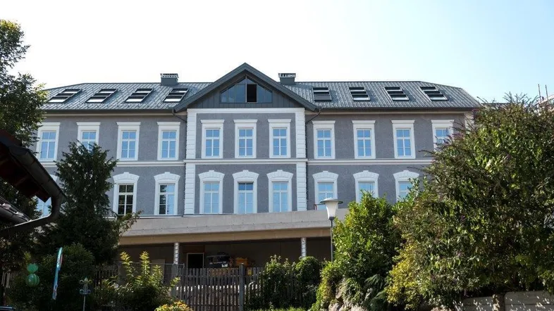 Klassisches Mehrfamilienhaus mit grauer Fassade, weißen Fensterrahmen und Dachfenstern unter blauem Himmel.