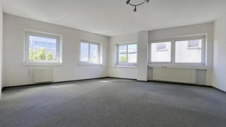 Geräumiger, leerer Büroraum mit grauem Teppichboden und mehreren Fenstern für viel Tageslicht.