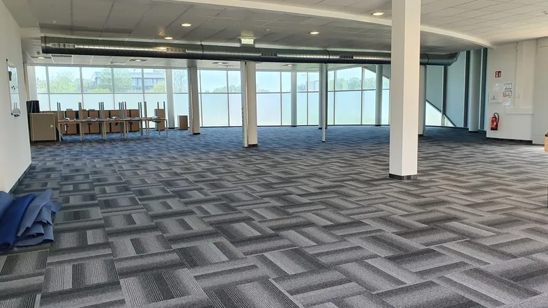 Großer, offener Bürobereich mit gemustertem Teppichboden, vielen Fenstern und sichtbaren Lüftungsrohren.