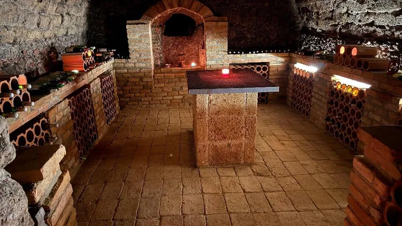 Historischer Weinkeller mit Ziegelgewölbe, Flaschenregalen und zentralem Steintisch, stimmungsvoll beleuchtet.